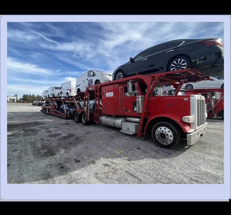 A red car carrier truck loaded with several vehicles parked on a lot under a partly cloudy sky.