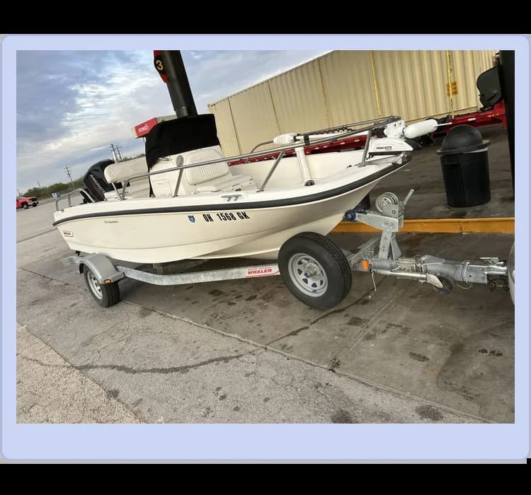 A small white motorboat on a trailer parked at a gas station with storage containers in the background under a cloudy sky.