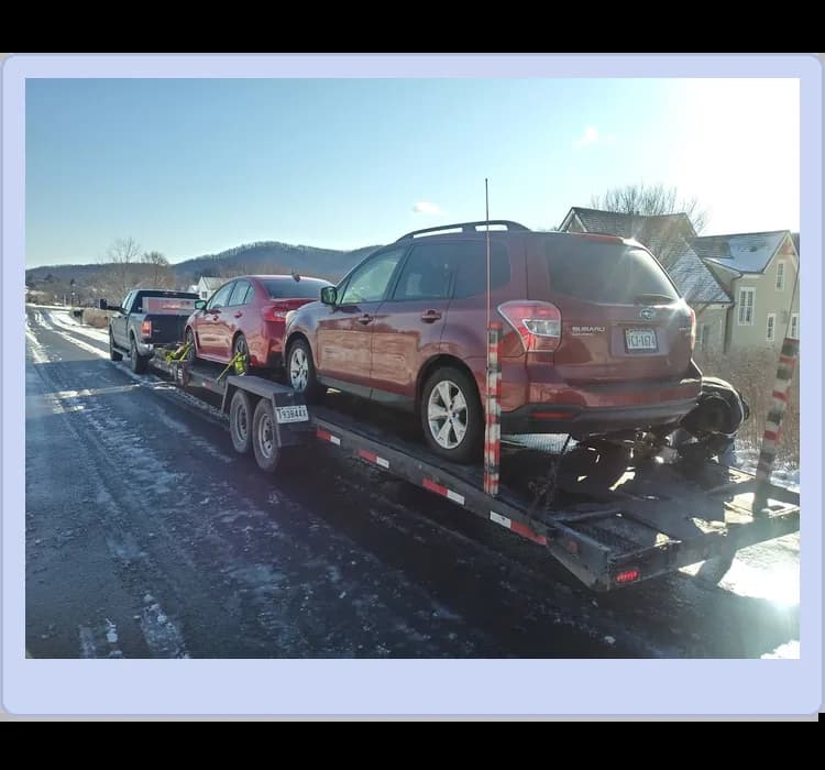 Two cars are loaded on a trailer being towed by a pickup truck on a snowy road with houses and bare trees in the background.