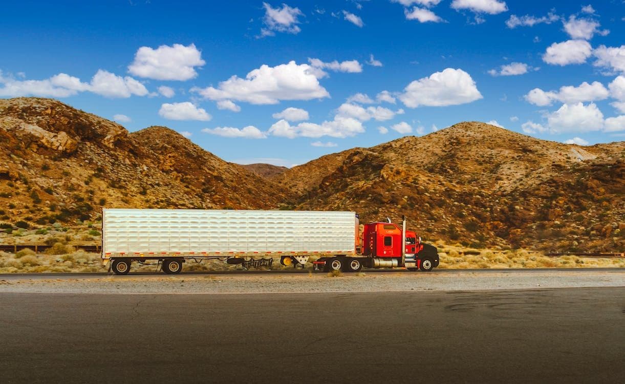 A red semi-truck with a white trailer drives along a road with mountains in the background under a blue sky.
