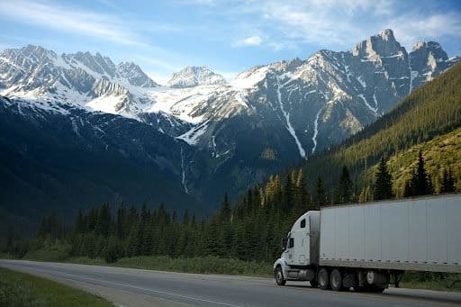 Semi-truck driving on a highway with snow-capped mountains in the background.
