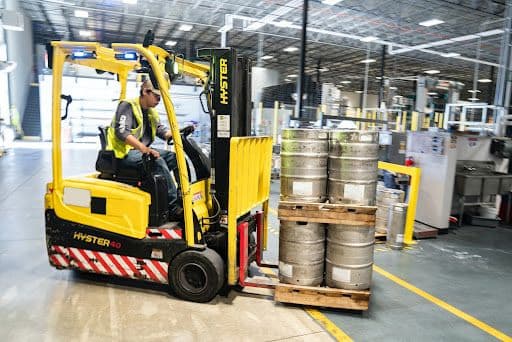 Worker operating a forklift to transport metal kegs in an industrial warehouse.