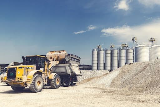 A wheel loader at a construction site moving gravel with silos in the background.