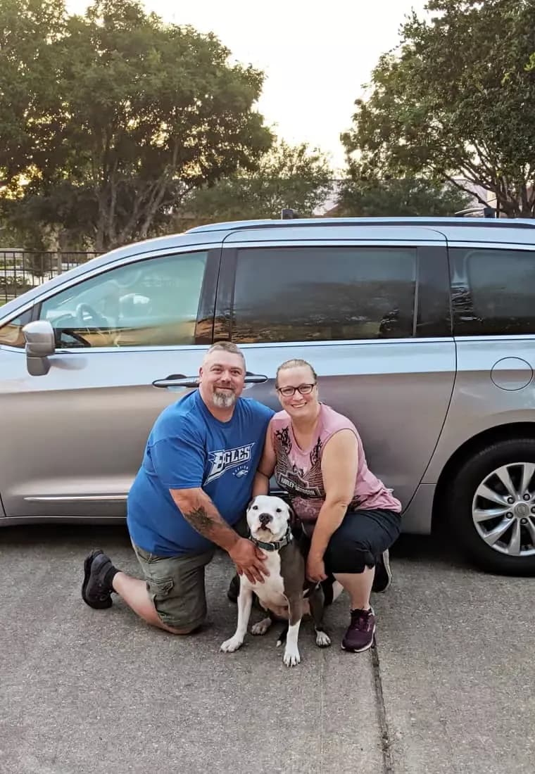 A couple posing happily with their dog in front of a silver minivan.
