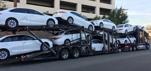 A car carrier truck transports multiple white sedans stacked in two rows on an open trailer, parked on a city street beside a large building.