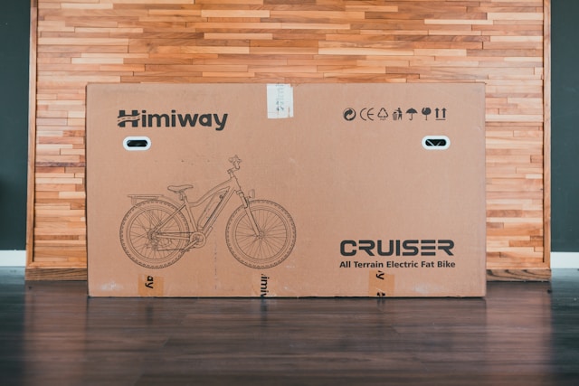 A cardboard box of a Himiway Cruiser electric fat bike sits against a wood-paneled wall, showcasing a line drawing of the bicycle and product details. This robust shipping package ensures your ride arrives safely and affordably.