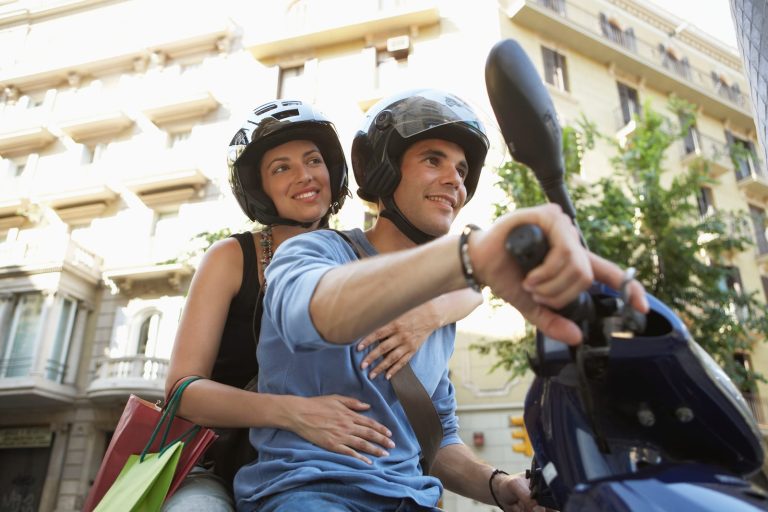 A man and woman wearing helmets ride an electric scooter through the city streets, with the woman holding shopping bags.