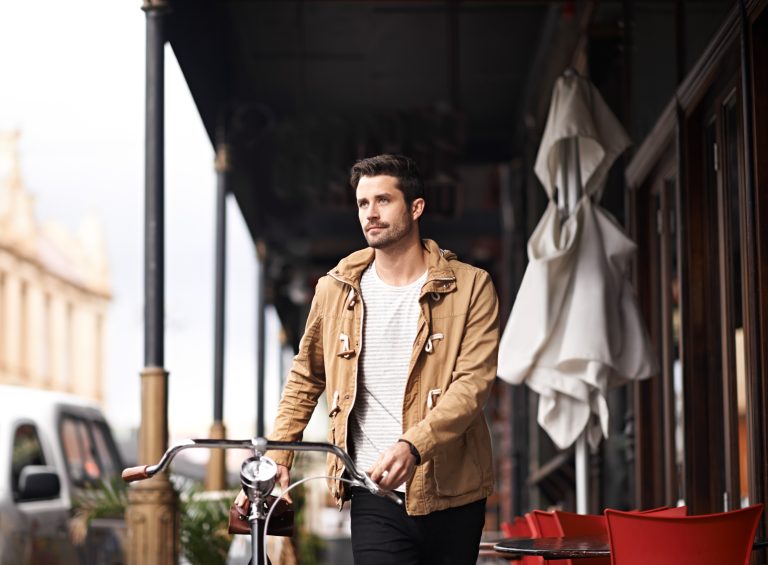 A man in a brown jacket walks beside his cheap bicycle on a city street, passing outdoor seating with red chairs and a cafe.