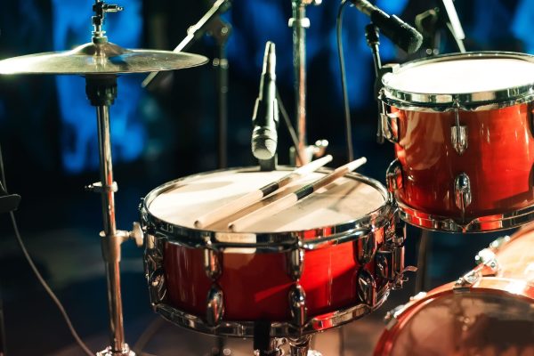 A red shipping drum set with drumsticks resting on the snare, surrounded by microphones, under vibrant stage lighting.