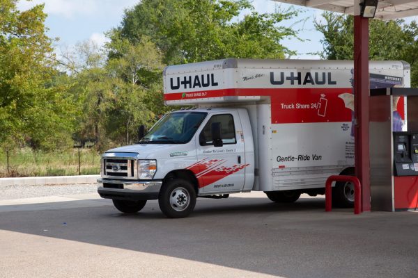 A U-Haul moving truck with its shipping doors closed is parked at a gas station near a fuel pump on a sunny day, with trees visible in the background.