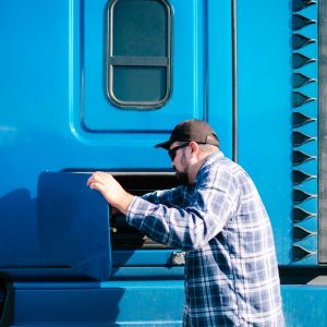 In the world of hot shot trucking, a man in a plaid shirt and cap expertly opens a compartment on his trusty blue truck.