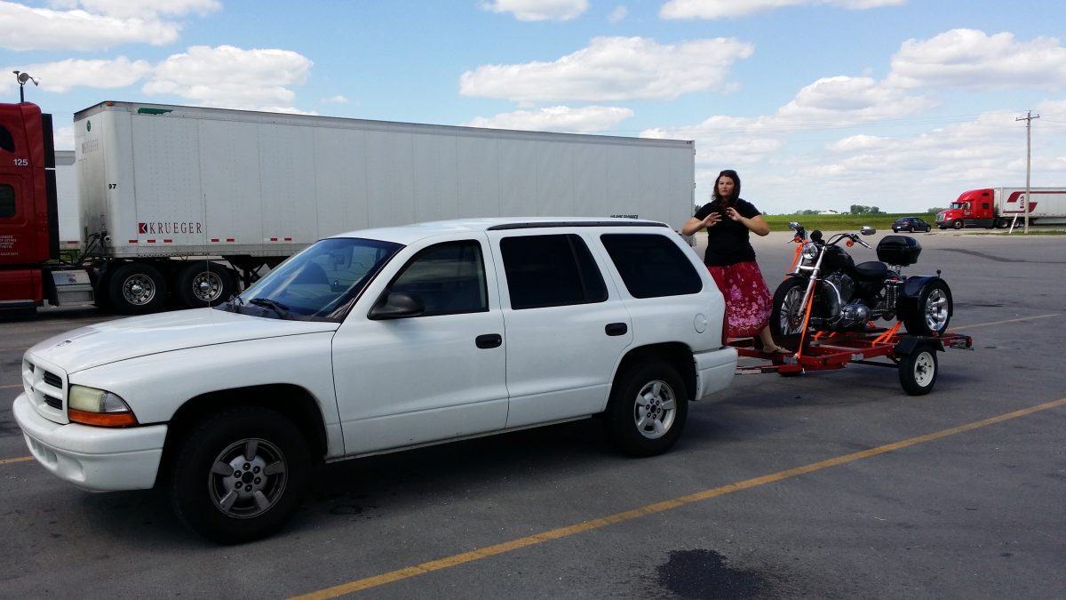 A white SUV, marked with a CitizenShipper decal, is parked in a lot towing a motorcycle on a small trailer. A woman stands behind the trailer, holding her phone. Semi-trucks and a Driver of the Month banner are visible in the background.
