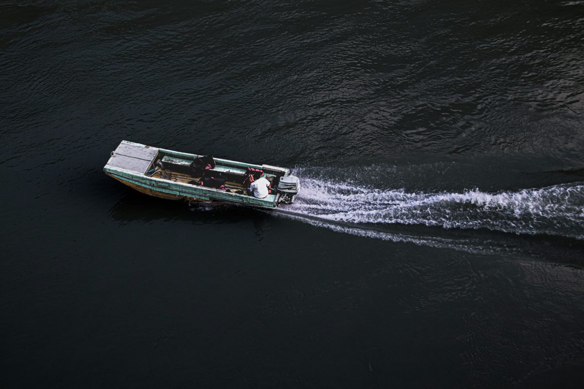 A small motorboat with two people moves quickly across dark water, leaving a visible wake behind—perfect for those interested in how to ship a powerboat or learn more about Powerboat Shipping.