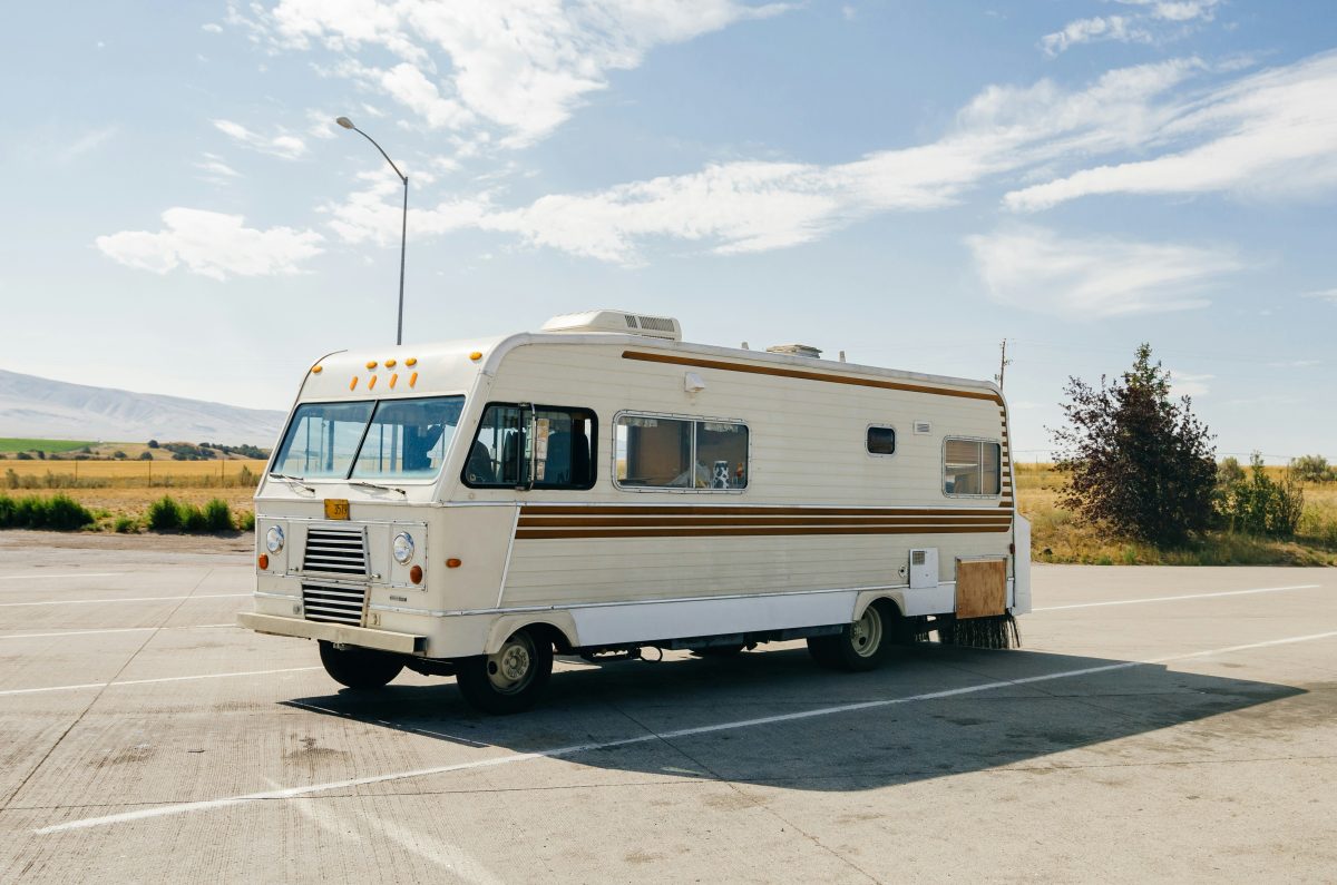 A vintage white and beige RV is parked in an empty lot on a sunny day with clear skies and open fields—perfect scenery for those curious about how to ship an RV or looking into RV trailer shipping options.