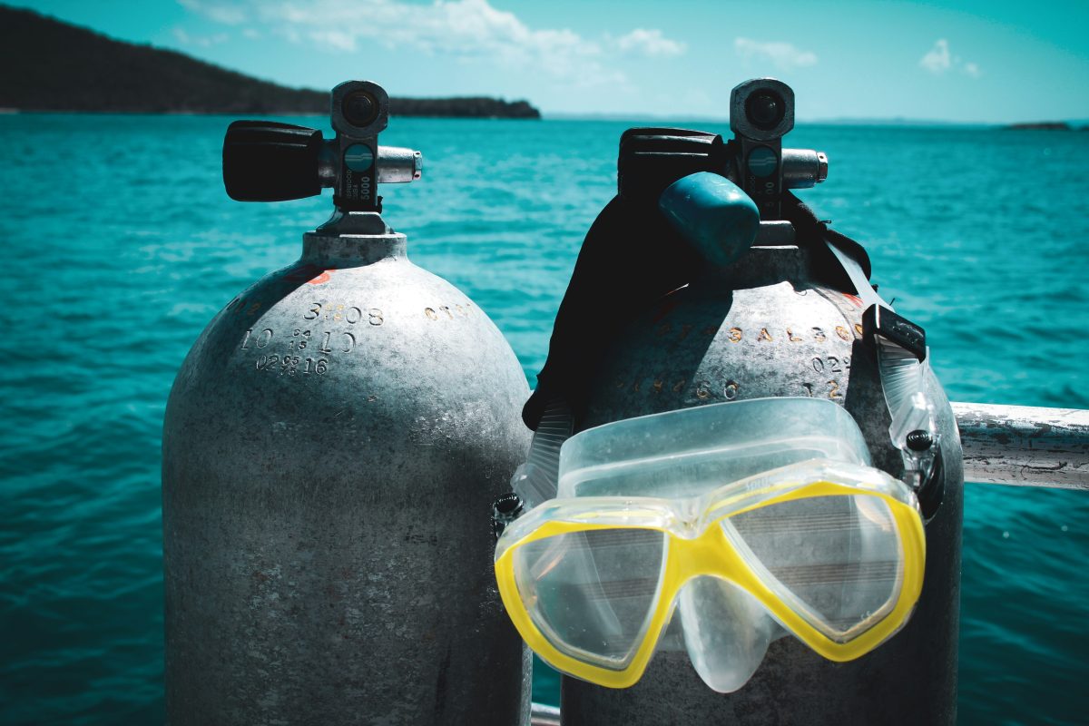 Two scuba tanks stand side by side on a boat with a diving mask draped over one, overlooking a blue ocean under a partly cloudy sky—perfect inspiration for learning how to ship scuba gear safely and efficiently.