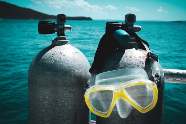 Two scuba tanks stand side by side on a boat with a diving mask draped over one, overlooking a blue ocean under a partly cloudy sky—perfect inspiration for learning how to ship scuba gear safely and efficiently.