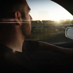 A man drives a car on the road at dusk, looking forward. Sunlight and reflections are visible through the window, partially illuminating his face.