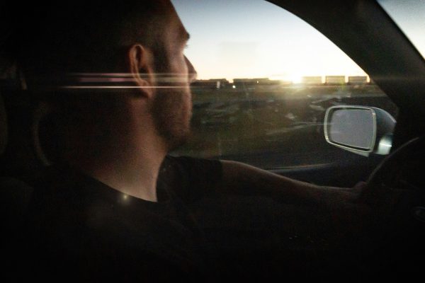 A man drives a car on the road at dusk, looking forward. Sunlight and reflections are visible through the window, partially illuminating his face.