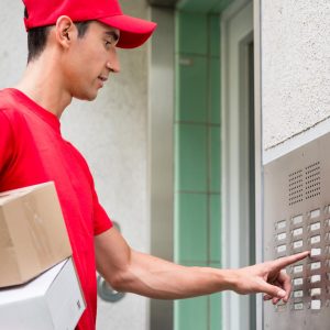 A delivery person in a red cap and shirt, specializing in residential freight shipping, holds packages while pressing a button on an intercom panel outside a building.