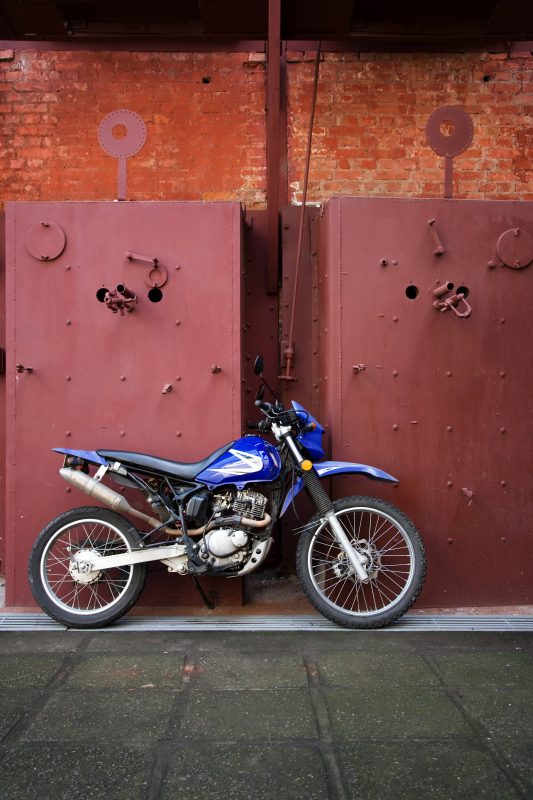 A blue dirt bike parked with motorcycle hitch carriers in front of a red door.