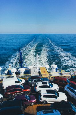 Cars parked on the deck of a ferry from a Car Transport Company glide across the sea, leaving a trail in the water under a clear blue sky.