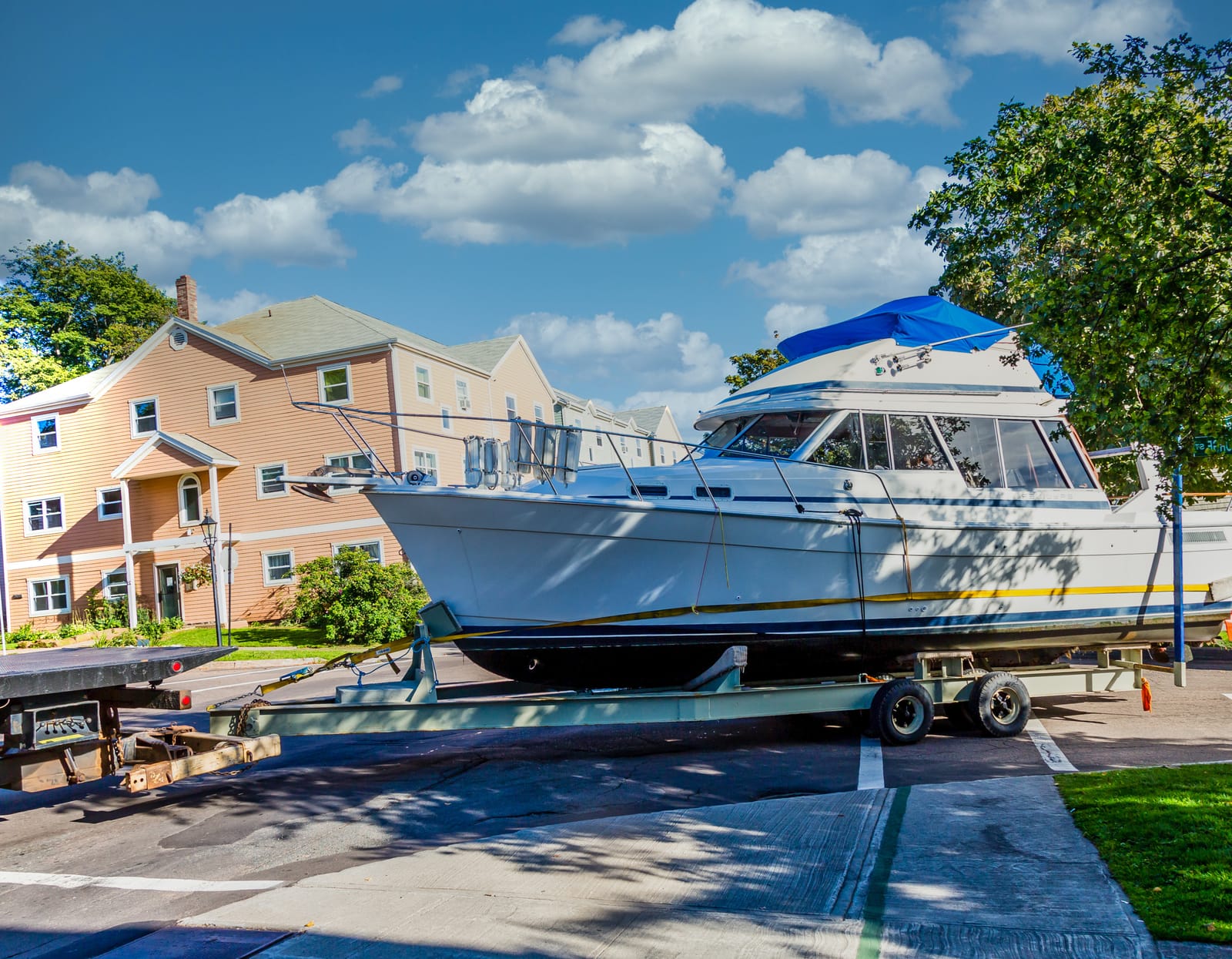 A large boat on a trailer, illustrating how to move a boat, is parked on a residential street near beige apartment buildings under a partly cloudy sky.