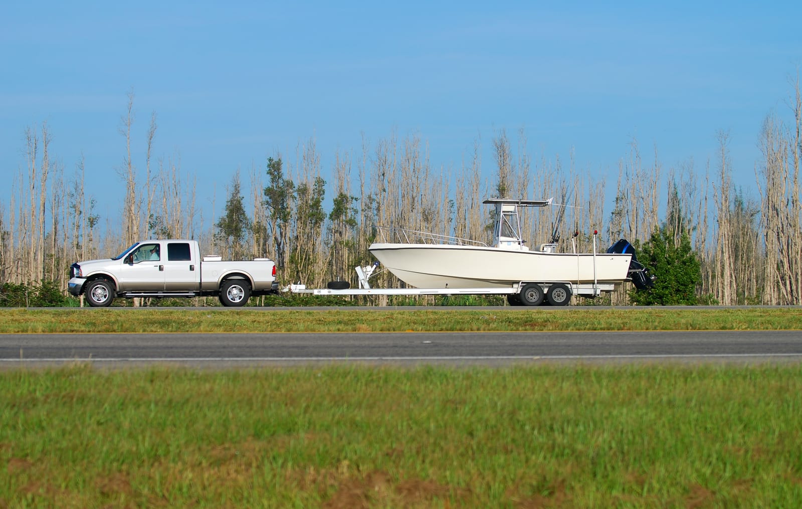 A white pickup truck is towing a large white boat on a trailer along a road with trees in the background, demonstrating how to move a boat safely and efficiently.