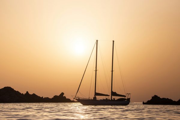 Silhouette of a sailboat gliding smoothly on calm water at sunset, showcasing the grace of sailboat transport. The clear sky and sun cast a warm glow, while rocks form a serene backdrop.