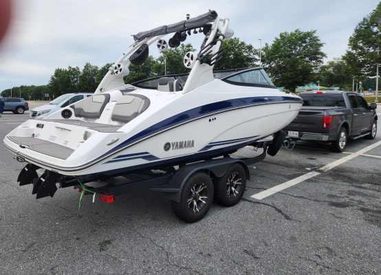 A white Yamaha speedboat with black and blue accents is parked on a trailer, hitched to a dark gray pickup truck—ready for transport by Convoy Commander on CitizenShipper—with trees and a cloudy sky in the background.