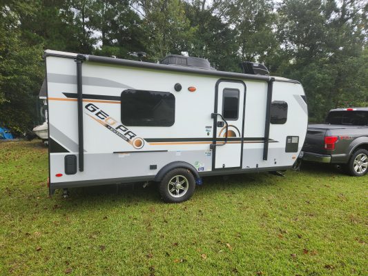 A small white and gray travel trailer labeled "Geo Pro" is parked on grass beside a gray pickup truck, as if awaiting its next CitizenShipper journey. Trees provide a peaceful backdrop for this travel-ready scene.