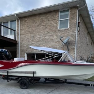 A vintage red and white motorboat with a canopy is parked on a trailer in front of a two-story brick house with large windows and a satellite dish, ready for transport by CitizenShipper’s trusted Convoy Commander.