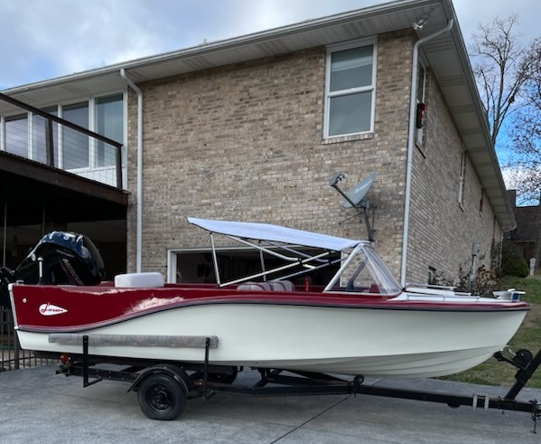 A vintage red and white motorboat with a canopy is parked on a trailer in front of a two-story brick house with large windows and a satellite dish, ready for transport by CitizenShipper’s trusted Convoy Commander.