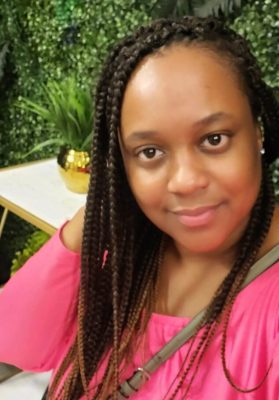 A woman with long braided hair, wearing a pink off-the-shoulder top, smiles at the camera. Seated in front of a leafy green wall and a table with a potted plant, she appears ready for her transporter interview.