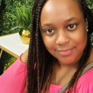 A woman with long braided hair, wearing a pink off-the-shoulder top, smiles at the camera. Seated in front of a leafy green wall and a table with a potted plant, she appears ready for her transporter interview.