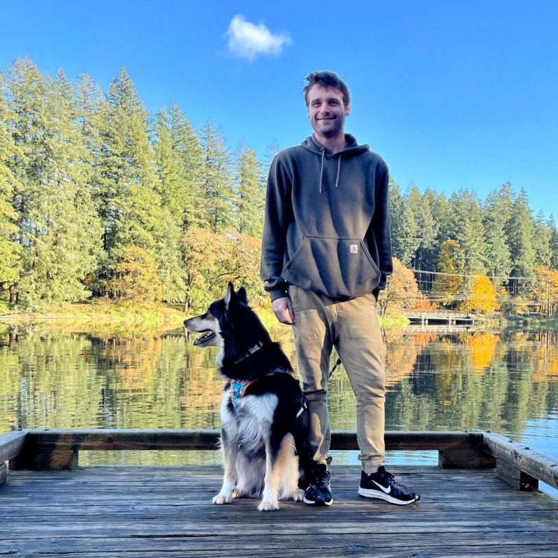 A man in a gray hoodie stands on a wooden dock with his black and white dog, ready for a transporter interview by the calm lake. Trees surround them under a blue sky with a single cloud.