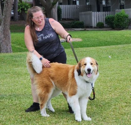 A woman with long hair and glasses, wearing a tank top and shorts, stands on grass holding the leash of a large, fluffy brown-and-white dog; together they share heartfelt journeys as CitizenShipper driver Julie Ann Jakubowski smiles at the camera.