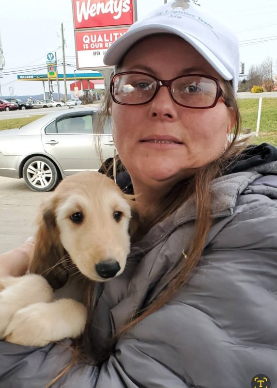 CitizenShipper driver Julie Ann Jakubowski, wearing glasses, a white hat, and a gray jacket, lovingly holds a light-colored puppy outside near parked cars, with a Wendy’s restaurant sign visible in the background—a moment from her heartfelt journeys.