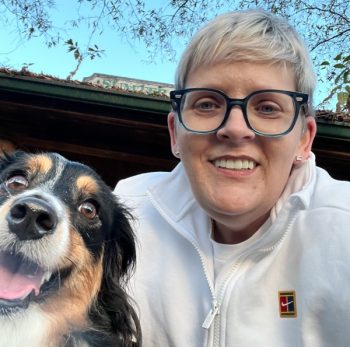 A woman with short blonde hair and glasses takes a selfie with a black and tan dog, both smiling at the camera, capturing their Journey to Happiness.