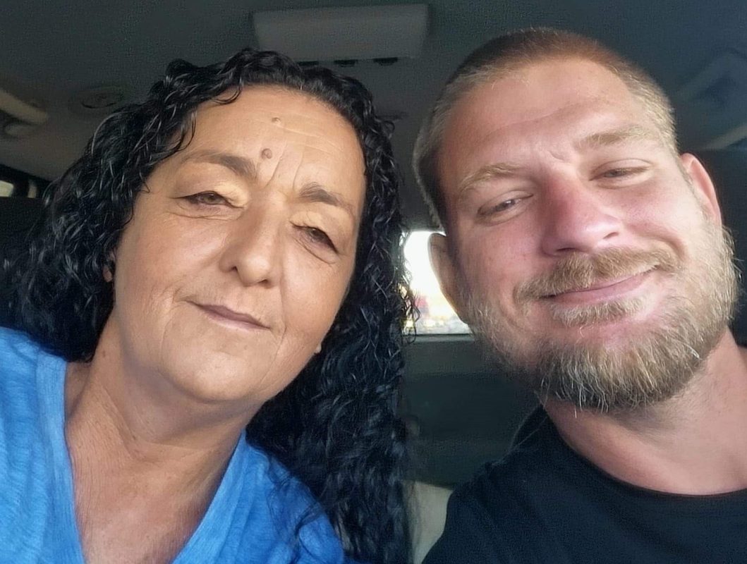 A woman with long curly hair and a blue shirt sits next to a man with short hair and a beard, both smiling at the camera inside a Noah's Ark Transport vehicle.