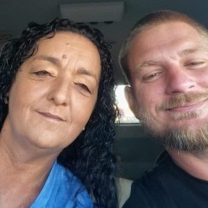 A woman with long curly hair and a blue shirt sits next to a man with short hair and a beard, both smiling at the camera inside a Noah's Ark Transport vehicle.