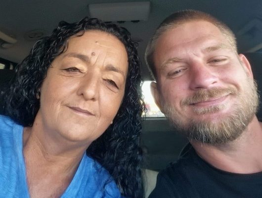 A woman with long curly hair and a blue shirt sits next to a man with short hair and a beard, both smiling at the camera inside a Noah's Ark Transport vehicle.