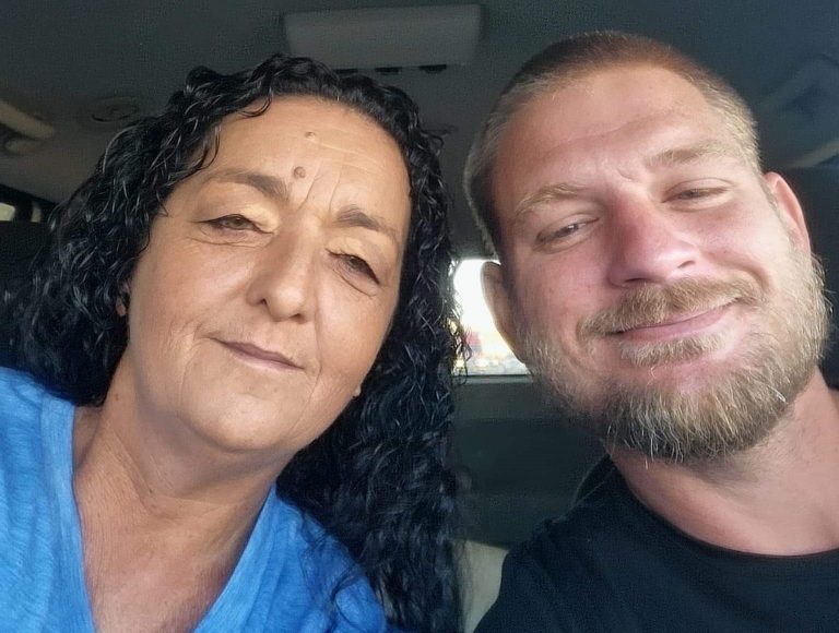 A woman with long curly hair and a blue shirt sits next to a man with short hair and a beard, both smiling at the camera inside a Noah's Ark Transport vehicle.