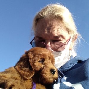 An older person with light hair, glasses, and a face mask holds a fluffy brown puppy close to their chest under a clear blue sky, perhaps after a heartwarming transporter interview.
