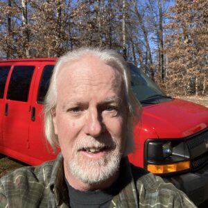 Phil Berry, a marine veteran with white hair and beard, is smiling proudly in front of his red Chevrolet van parked in a wooded area. Now a dedicated pet transporter, he finds joy in bringing furry friends safely to their new homes.