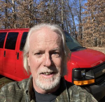 Phil Berry, a marine veteran with white hair and beard, is smiling proudly in front of his red Chevrolet van parked in a wooded area. Now a dedicated pet transporter, he finds joy in bringing furry friends safely to their new homes.