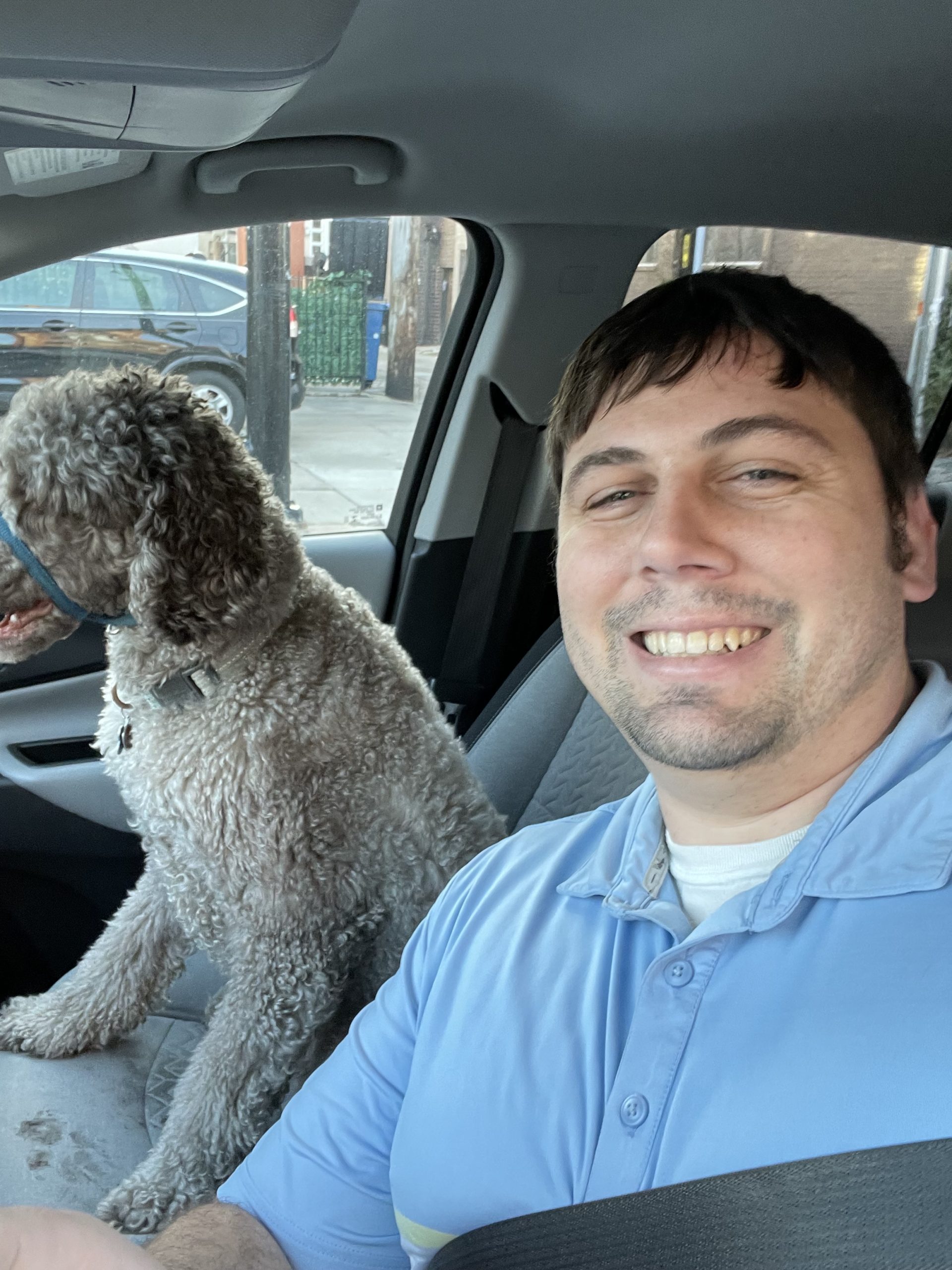 A CitizenShipper driver smiles in a car while a curly-haired dog sits in the passenger seat, eagerly looking out the window.