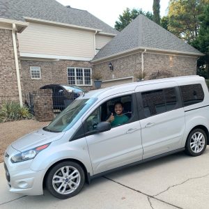A person smiles and gives a thumbs-up while sitting in the driver's seat of a silver van, ready for compassionate and safe pet transport. Trees, a garage, and a brick house are visible in the background.