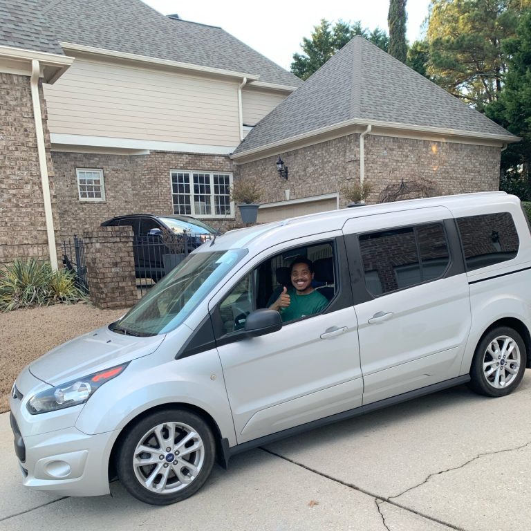 A person smiles and gives a thumbs-up while sitting in the driver's seat of a silver van, ready for compassionate and safe pet transport. Trees, a garage, and a brick house are visible in the background.