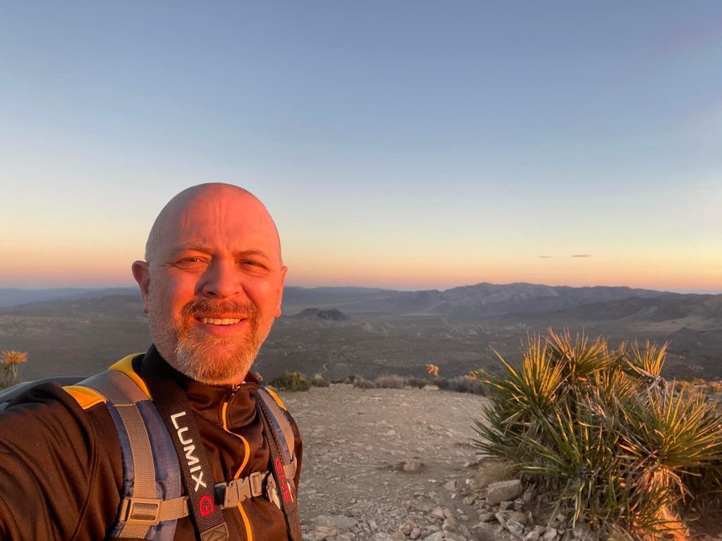 A journey unfolds as a person in hiking gear stands on a rocky path at sunset, with a vast desert landscape and distant mountains stretching endlessly in the background.