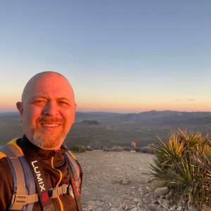 A journey unfolds as a person in hiking gear stands on a rocky path at sunset, with a vast desert landscape and distant mountains stretching endlessly in the background.